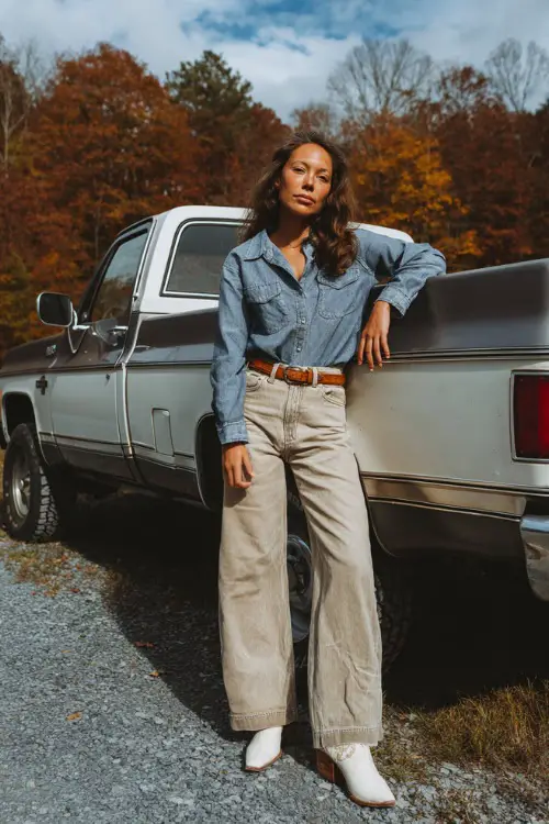 A woman in wide-leg jeans, tucked chambray shirt, tan belt, and white cowgirl boots, leaning casually against a pickup truck with autumn trees in the background