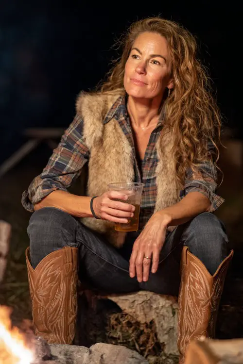 A woman over 40 styled in dark flared jeans, plaid button-down shirt, faux fur vest, and tan cowboy boots 