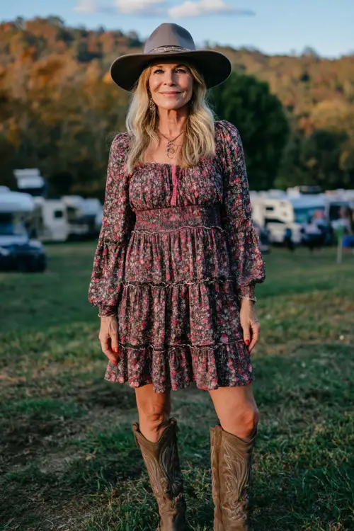 A woman over 40 wearing a tiered floral dress with long sleeves, accessorized with a felt hat and knee-high cowboy boots 
