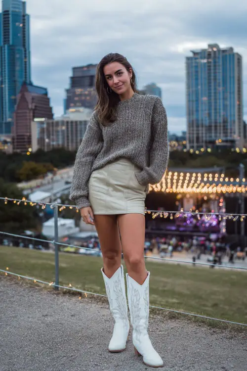 A woman styled in a leather mini skirt, tucked-in knit sweater, and tall white cowboy boots, standing near an outdoor concert stage under string lights