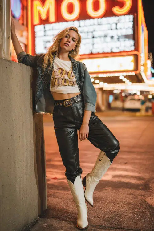 A woman styled in high-waisted black leather pants, tucked graphic tee, cropped denim jacket, and sleek white cowboy boots, posing near neon festival signs at night