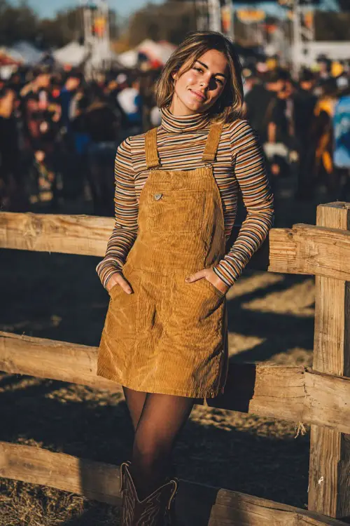 A woman wearing a corduroy pinafore dress layered over a striped turtleneck, paired with tights and cowgirl boots, standing by a wooden fence with a concert crowd behind her 