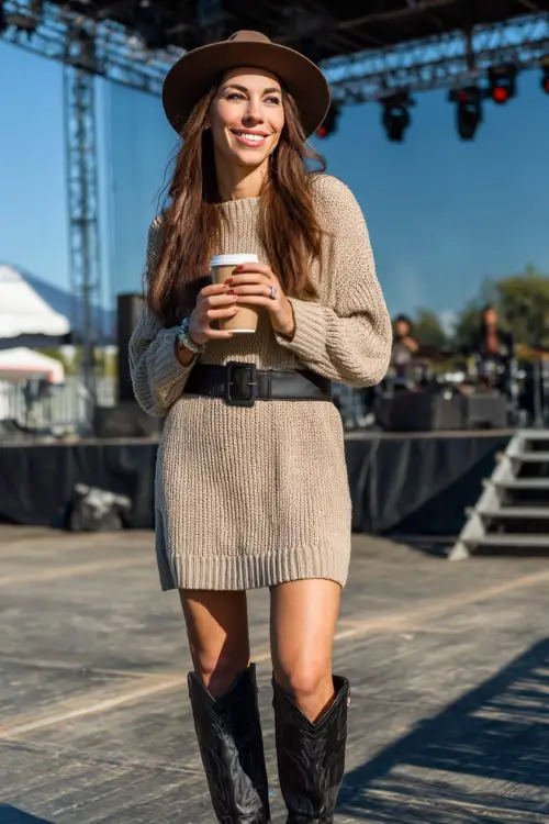 A woman wearing a long-sleeve knit sweater dress with a leather belt, paired with tall black cowboy boots and a wool fedora hat, holding a coffee near an outdoor concert stage 