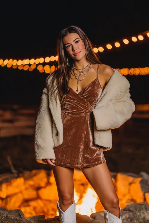 A woman wearing a velvet wrap dress with a cropped faux fur jacket, accessorized with layered necklaces, paired with sleek white cowboy boots, standing near a firepit with glowing lights 