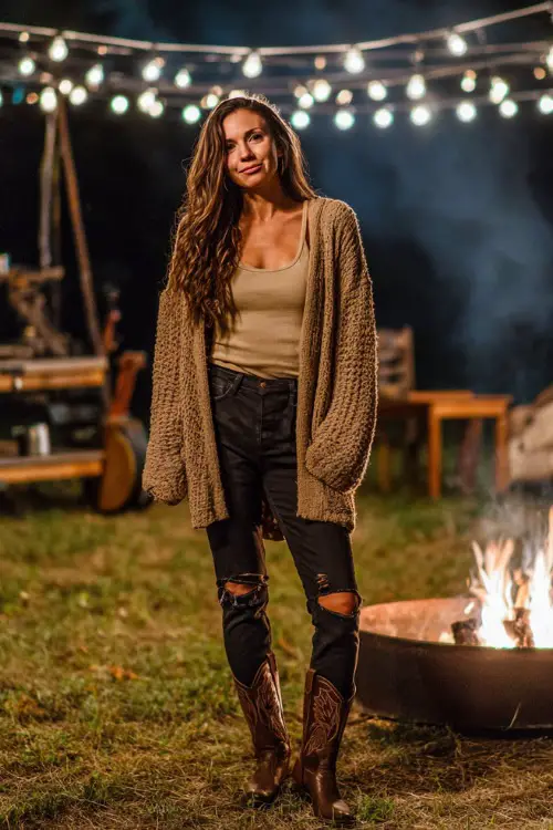 A woman wearing dark ripped jeans, a cozy oversized cardigan, tucked tank top, and cowgirl boots, standing near a firepit with glowing concert lights in the background