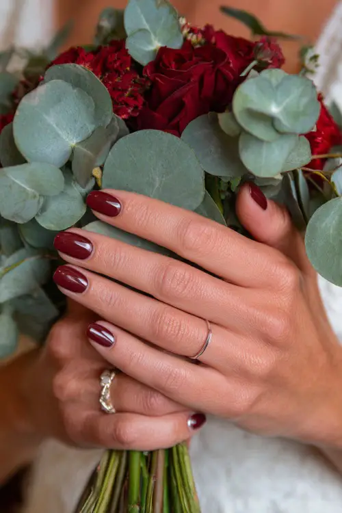 Bride’s hands with almond-shaped glossy burgundy nails, gently holding a bouquet of red roses and eucalyptus
