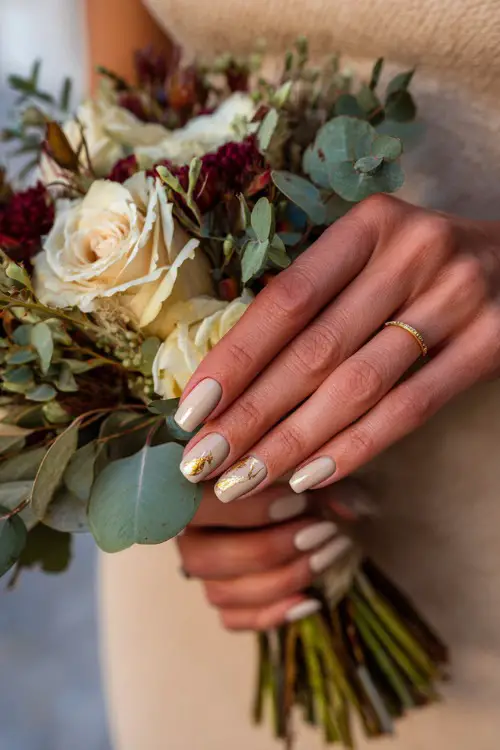 Bride’s hands with almond-shaped neutral beige nails decorated with delicate gold leaf accents, holding an autumn bridal bouquet of roses and eucalyptus 