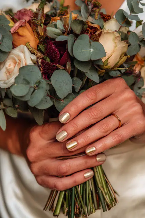 Bride’s hands with almond-shaped neutral beige nails decorated with delicate gold leaf accents, holding an autumn bridal bouquet of roses and eucalyptus