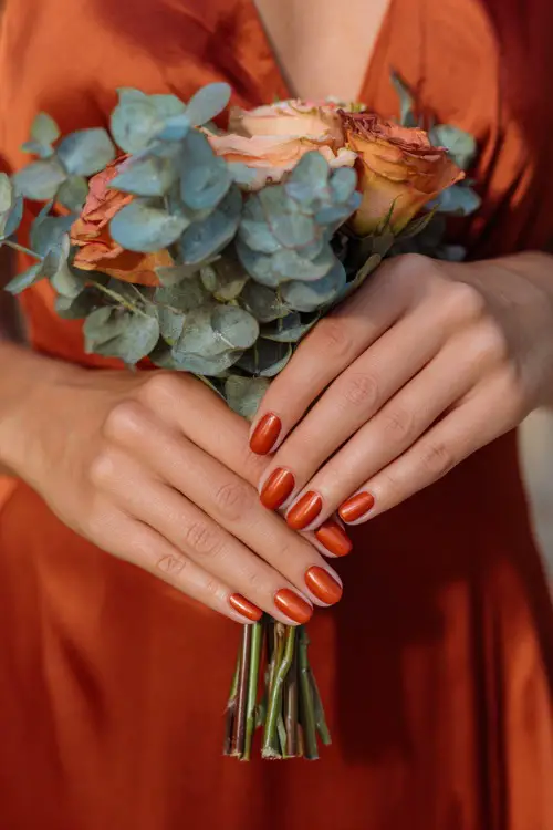 Bridesmaid’s hands with almond-shaped rust orange nails in glossy finish, holding a bouquet of fall roses and eucalyptus 