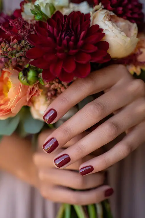 Bridesmaid’s hands with short glossy burgundy nails, holding a bouquet of dahlias and roses in fall colors
