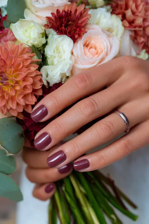 Bridesmaid’s hands with short glossy burgundy nails, holding a bouquet of dahlias and roses in fall colors