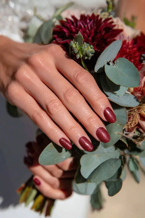 Burgundy nails with subtle copper glitter highlights, bride’s hands holding a fall bouquet of dahlias and eucalyptus 