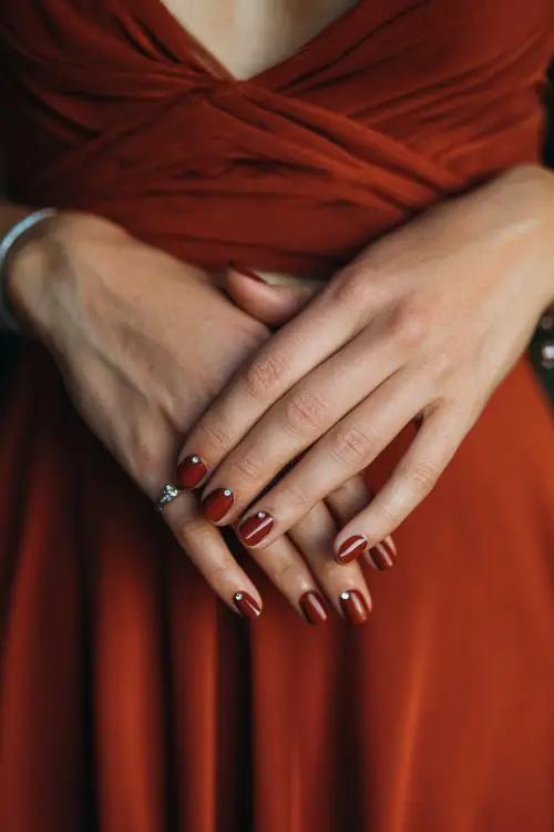 Dark rust matte nails with small rhinestone accents, bridesmaid’s hands placed over soft chiffon bridesmaid gown 