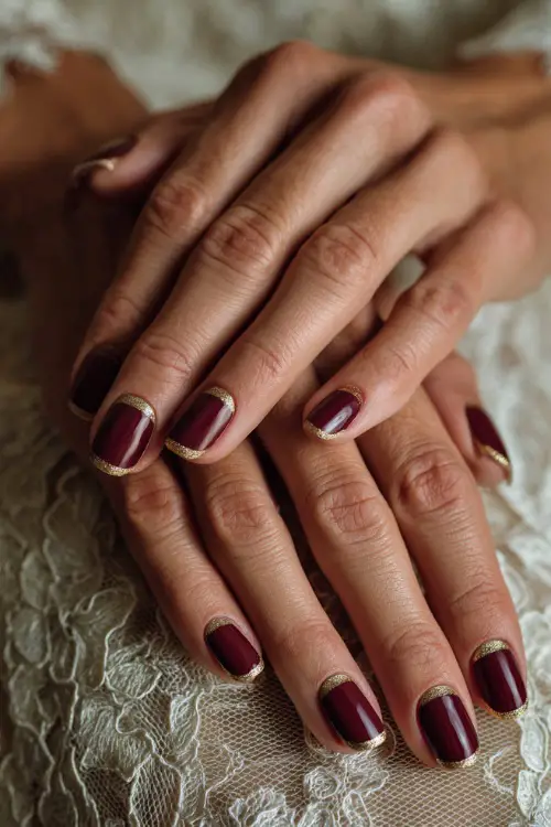 Elegant coffin-shaped burgundy nails with gold glitter tips, bride’s hands resting on lace wedding gown fabric 