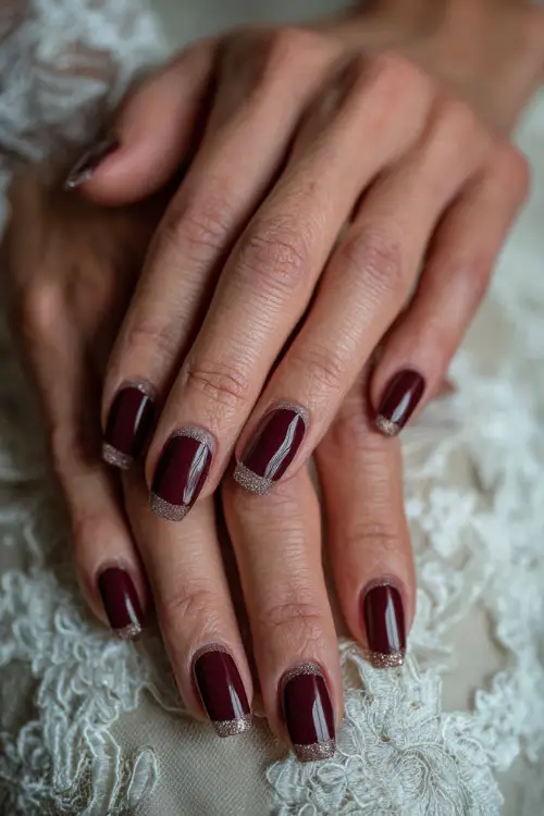 Elegant coffin-shaped burgundy nails with gold glitter tips, bride’s hands resting on lace wedding gown fabric (4)