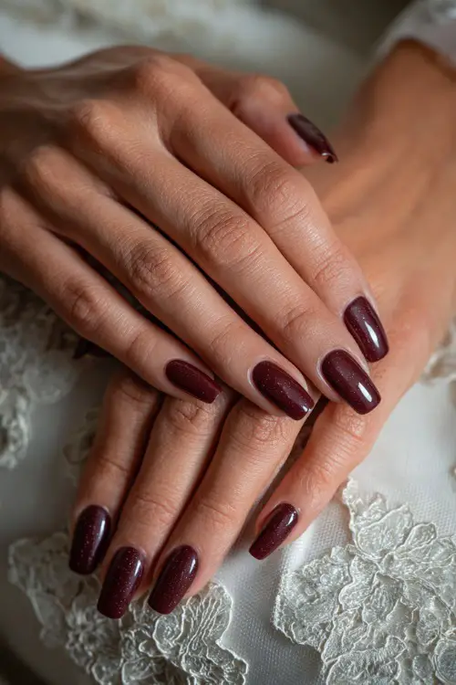 Elegant coffin-shaped burgundy nails with subtle glitter tips, bride’s hands resting on lace wedding dress fabric