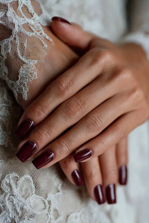 Elegant coffin-shaped burgundy nails with subtle glitter tips, bride’s hands resting on lace wedding dress fabric