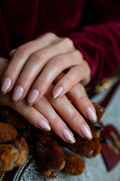 Elegant soft blush pink nails with gold glitter tips, guest’s hands resting on a clutch bag with autumn décor in the background
