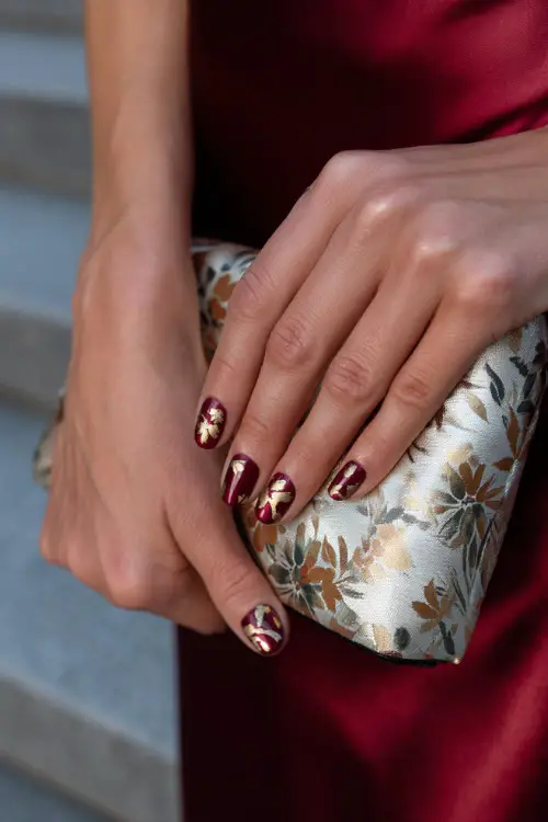 Glossy burgundy nails with hand-painted floral patterns in gold, bride’s hands holding a satin clutch 