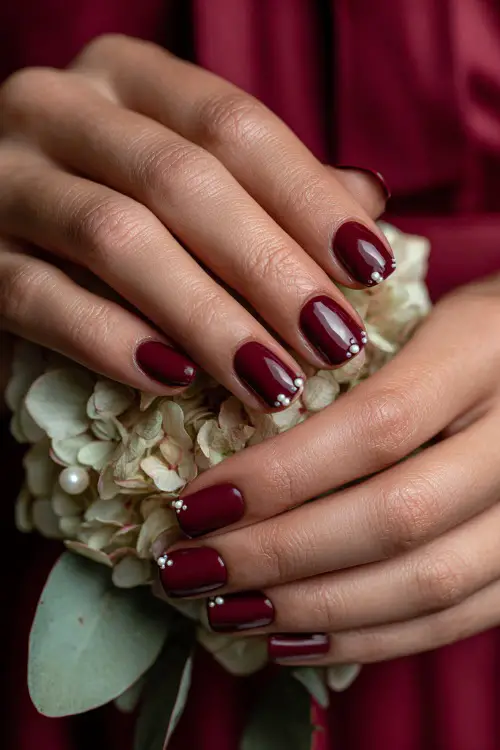 Glossy maroon nails with pearl-studded cuticle accents, bridesmaid’s hands holding a floral corsage 