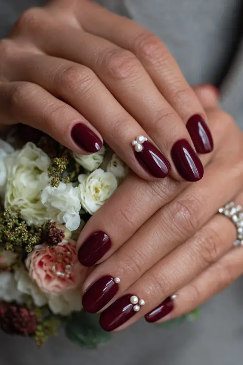 Glossy maroon nails with pearl-studded cuticle accents, bridesmaid’s hands holding a floral corsage 