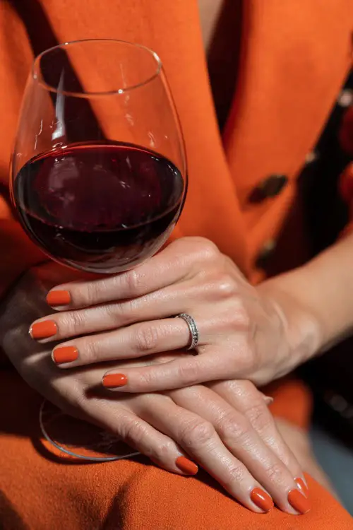 Guest’s hands with short almond-shaped burnt orange nails, holding a glass of red wine at a fall wedding reception