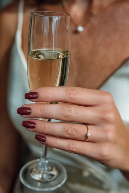 Matte burgundy nails with delicate pearl accents near the cuticle, bride holding a champagne flute at reception 