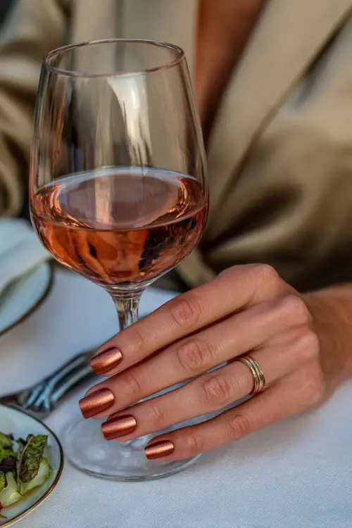 Rust-toned French manicure with copper metallic tips, bridesmaid’s hands lifting a wine glass at wedding dinner