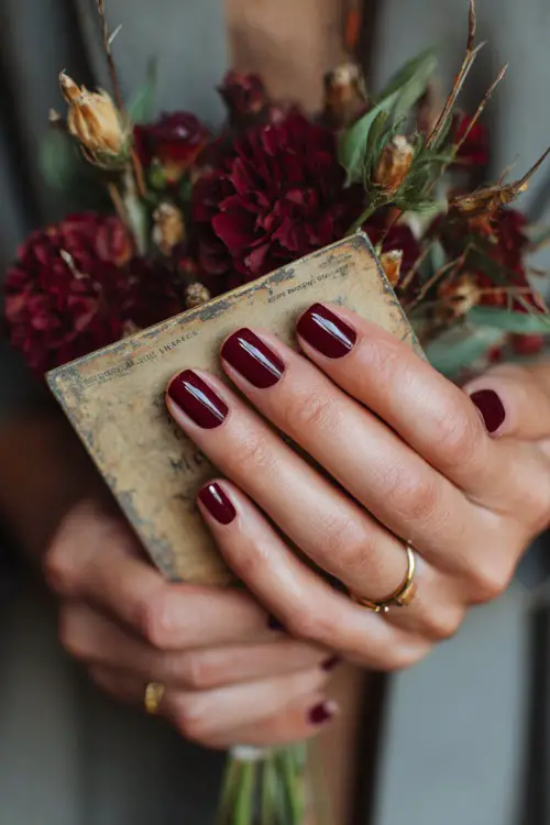 Short burgundy nails with fine gold foil accents, bride’s hands holding a rustic wedding invitation card 
