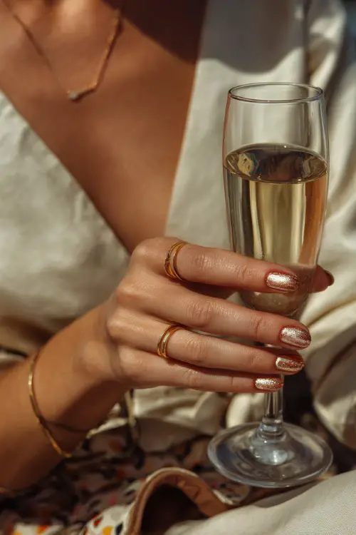 Short cream-colored nails with copper foil details, bridesmaid’s hand holding a glass of champagne 