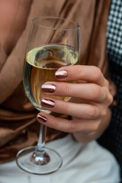 Short cream-colored nails with copper foil details, bridesmaid’s hand holding a glass of champagne