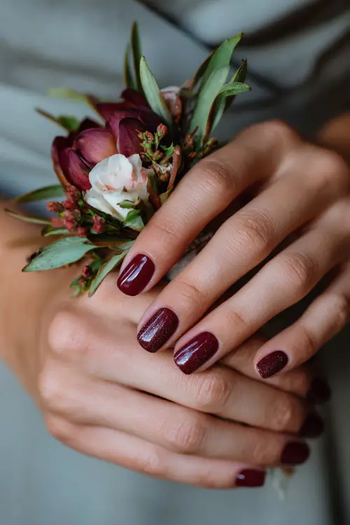 Short maroon nails with glitter ombré fade, bridesmaid’s hands gently holding a floral corsage