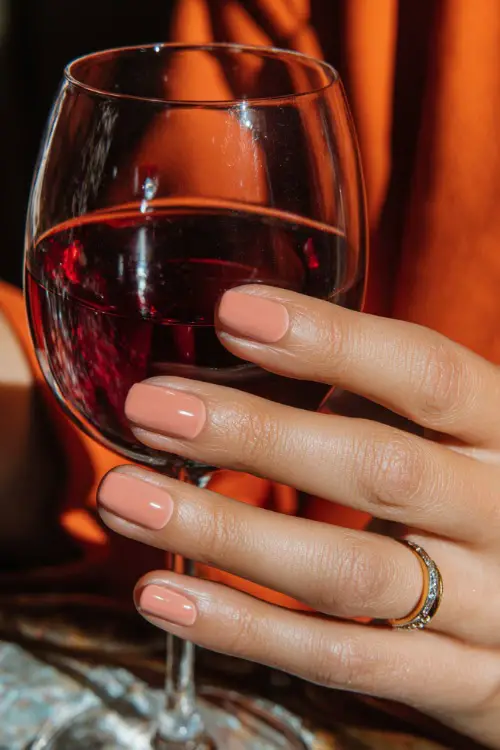 Short pale peach nails with glossy natural look, bridesmaid’s hand holding a glass of red wine at fall wedding dinner 