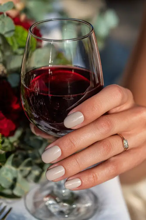 Short pale peach nails with glossy natural look, bridesmaid’s hand holding a glass of red wine at fall wedding dinner