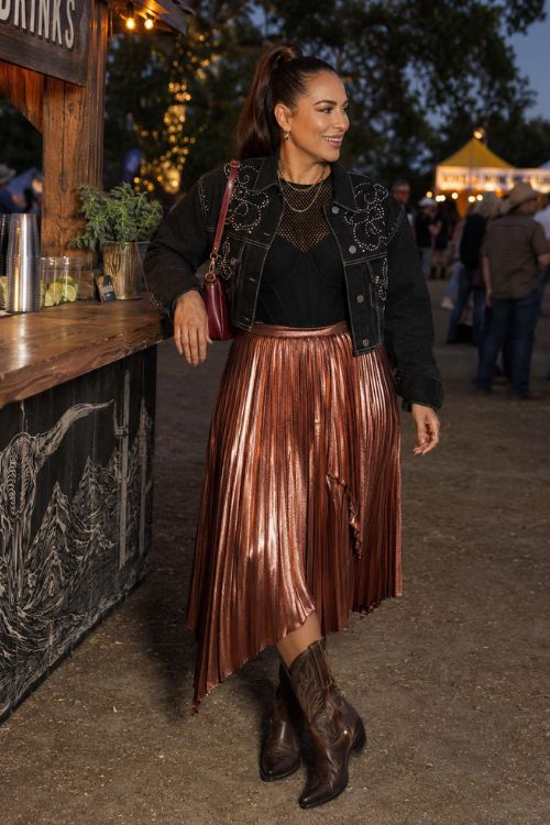 woman wearing a fall night country concert outfit with copper metallic pleated midi skirt, a fitted black mesh-knit top, a cropped dark denim jacket, and chocolate cowboy boots