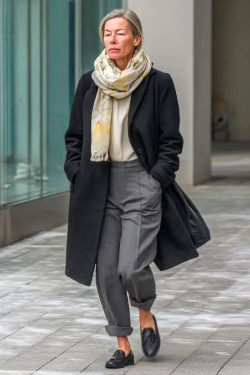 A woman in her 50s wears a black wool coat over a cream blouse and gray straight-leg trousers