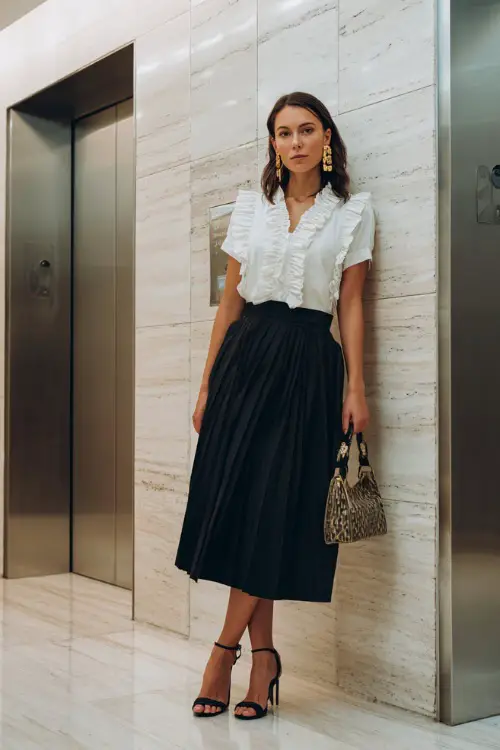 A woman wears a black pleated midi skirt with a white ruffled blouse and heeled sandals, accessorized with a small handbag and gold hoop earrings