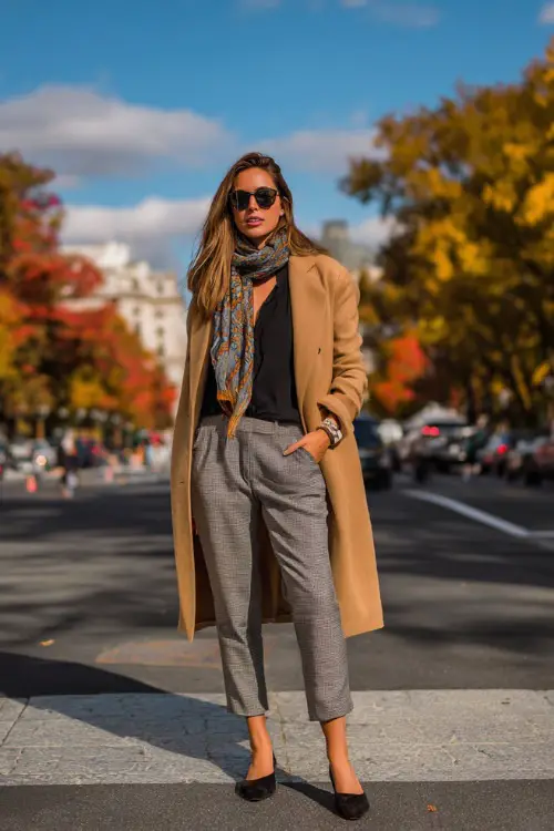 A woman wears a tan wool coat over a black blouse and gray trousers, accessorized with a scarf and heeled loafers