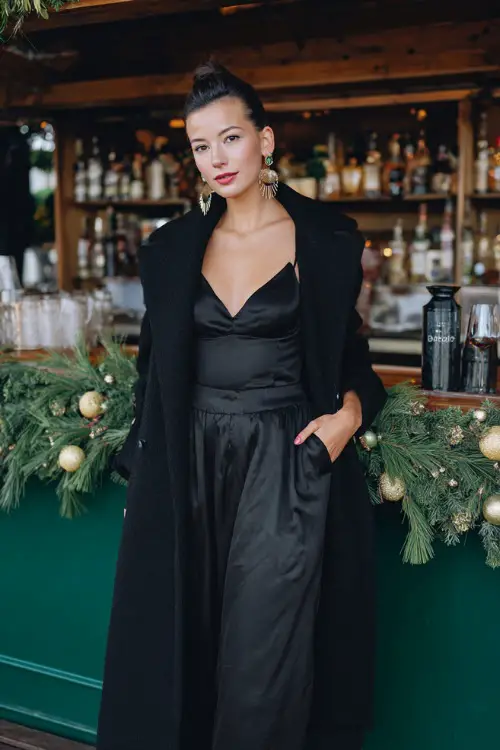 A woman in a black satin midi dress with a long tailored coat and bold earrings, standing near a holiday cocktail bar decorated with greenery and ornaments