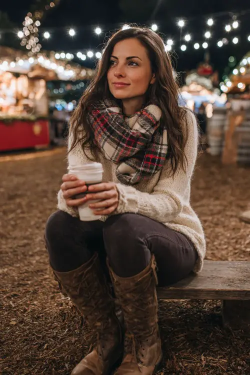 A woman in a cozy plaid scarf, cream sweater, and dark leggings with tall boots, enjoying a warm drink at an outdoor Christmas festival