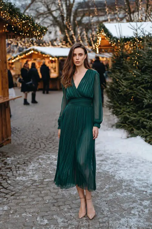 A woman in a forest-green pleated chiffon dress with long sleeves and ankle-strap heels, posing outside near a snowy Christmas market with warm twinkling lights
