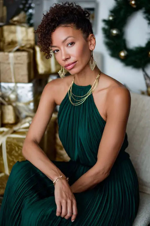A woman in an emerald green pleated dress with gold accessories, posing in a bright indoor studio filled with Christmas wreaths, gifts, and sparkling décor