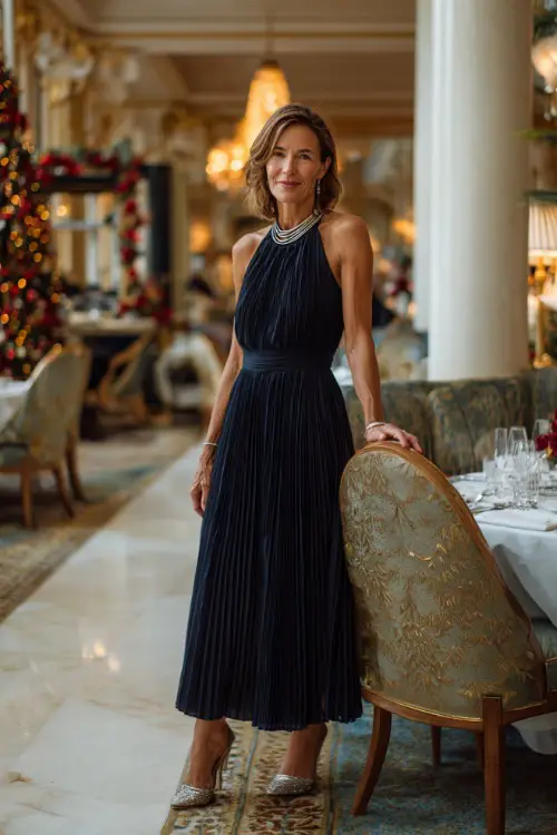A woman over 50 in a navy pleated midi dress with silver jewelry and elegant heels, posing inside a beautifully decorated Christmas dinner venue 
