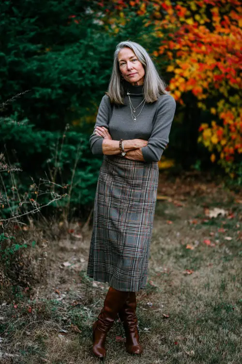 A woman over 50 in a plaid midi skirt paired with a turtleneck and knee-high boots, standing outdoors surrounded by colorful autumn trees 