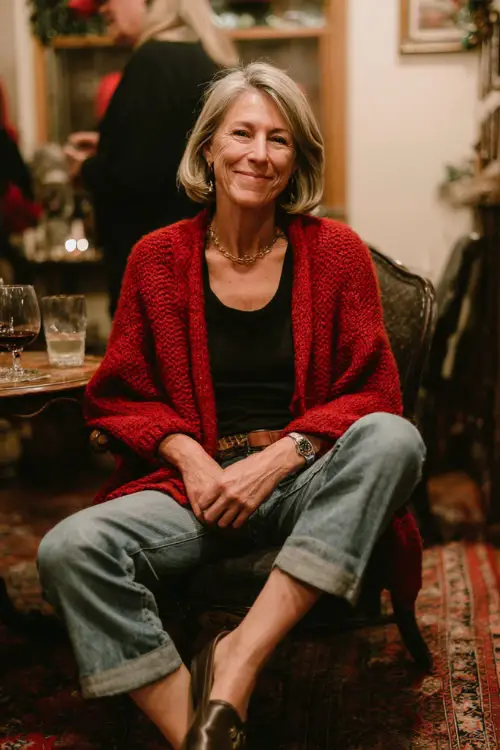 A woman over 50 in a red cardigan layered over a simple black top with straight-leg jeans and loafers, enjoying a relaxed holiday party with friends