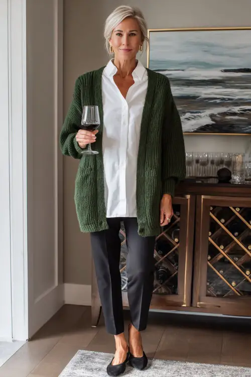 A woman over 50 wearing a forest-green cardigan over a white blouse with slim black pants and elegant flats, enjoying a cozy holiday home gathering