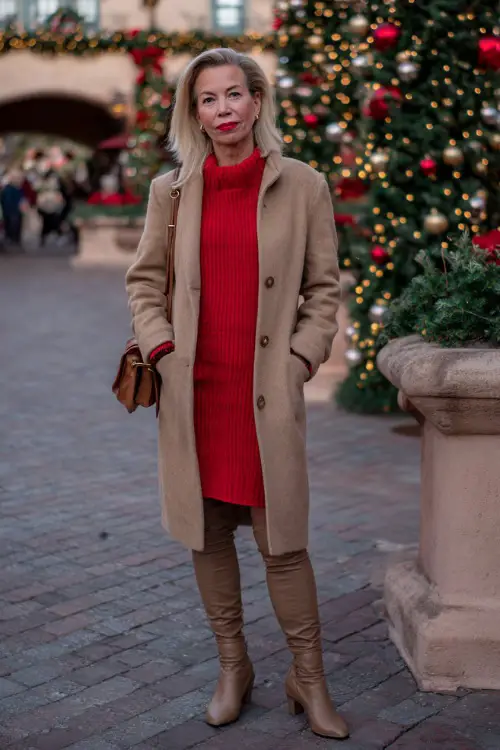 A woman over 50 wearing a tailored camel coat over a red sweater dress with tights and heeled boots