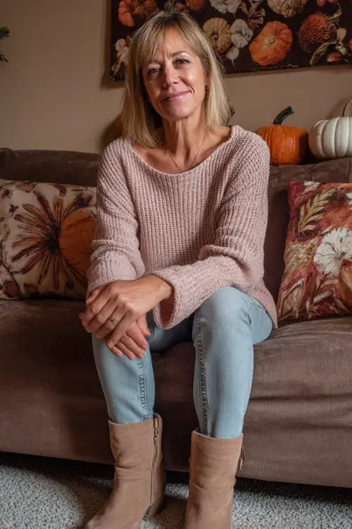 A woman over 50 wears a light pink knit pullover with straight jeans and suede ankle boots, sitting on a couch decorated with Thanksgiving pillows (