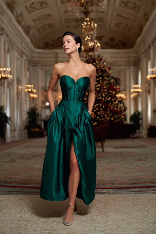 A woman wearing a deep emerald satin gown with a structured bodice, gold heels, and pearl drop earrings, standing in a luxurious holiday ballroom with chandeliers and a grand Christmas tree