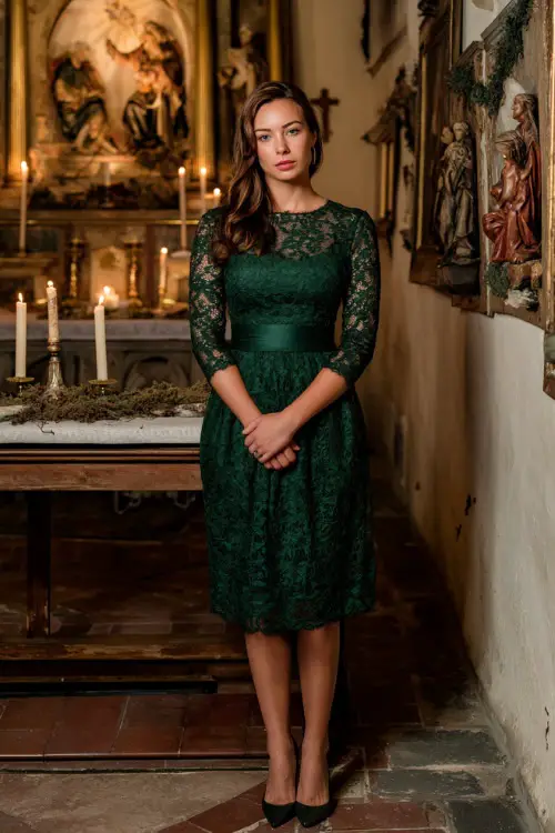 A woman wearing a modest emerald-green lace dress with long sleeves and classic pumps, standing in a church foyer decorated with nativity scenes and candles 